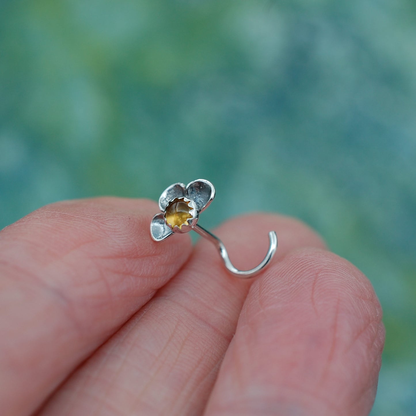 Silver flower-shaped nose stud with a citrine gemstone held between fingers against a blurred natural background