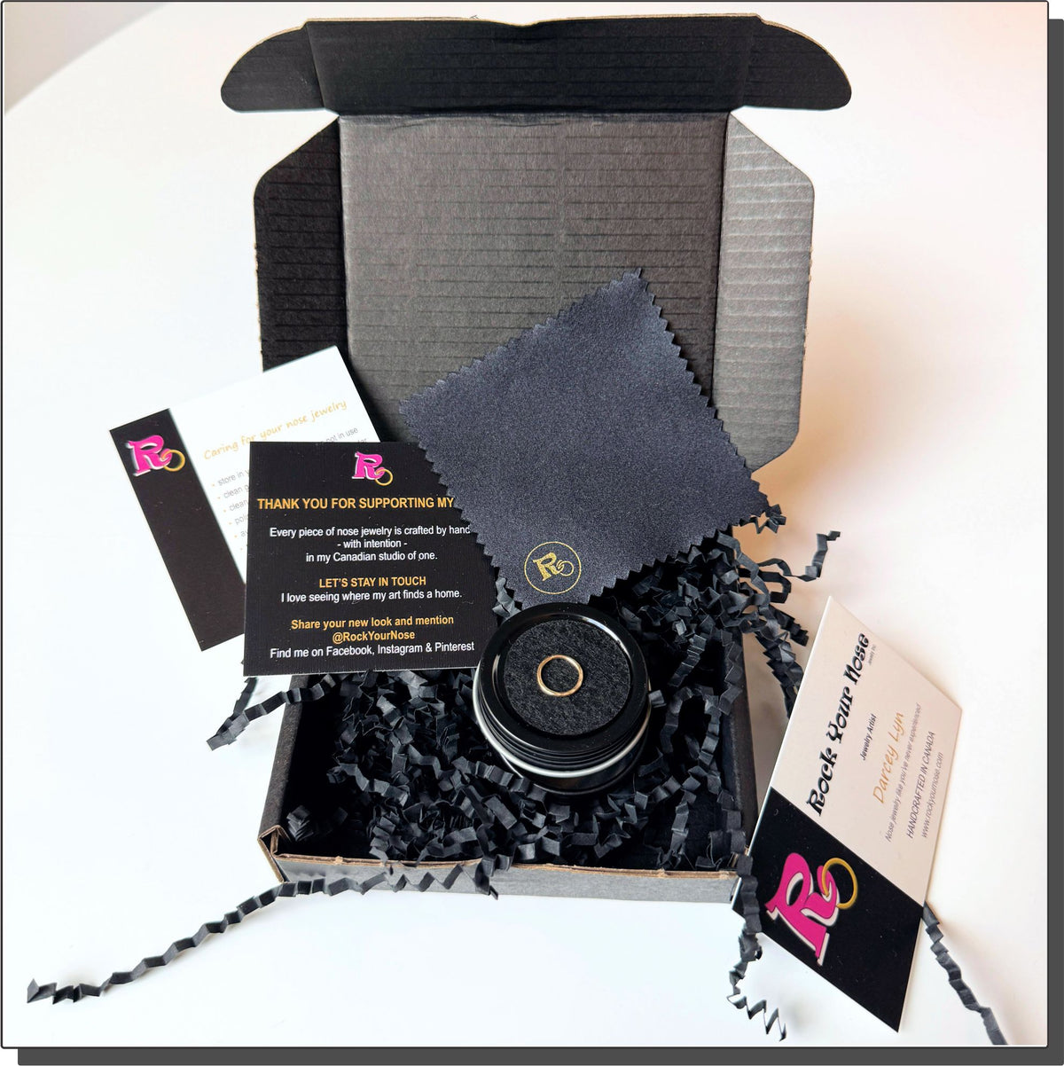 A premium black unboxing experience for Rock Your Nose handcrafted nose jewelry. An open matte-black cardboard mailer box filled with black crinkle paper cleanly showcases a circular black tin container displaying a single gold nose ring on a dark cushion. Elegant black-and-white insert cards are neatly layered on the right, displaying nose jewelry care instructions and a thank you message from the Canadian artisan studio of one.