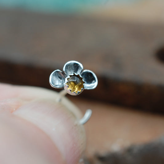 Silver flower-shaped nose stud with a yellow gemstone held between fingers against a blurred background