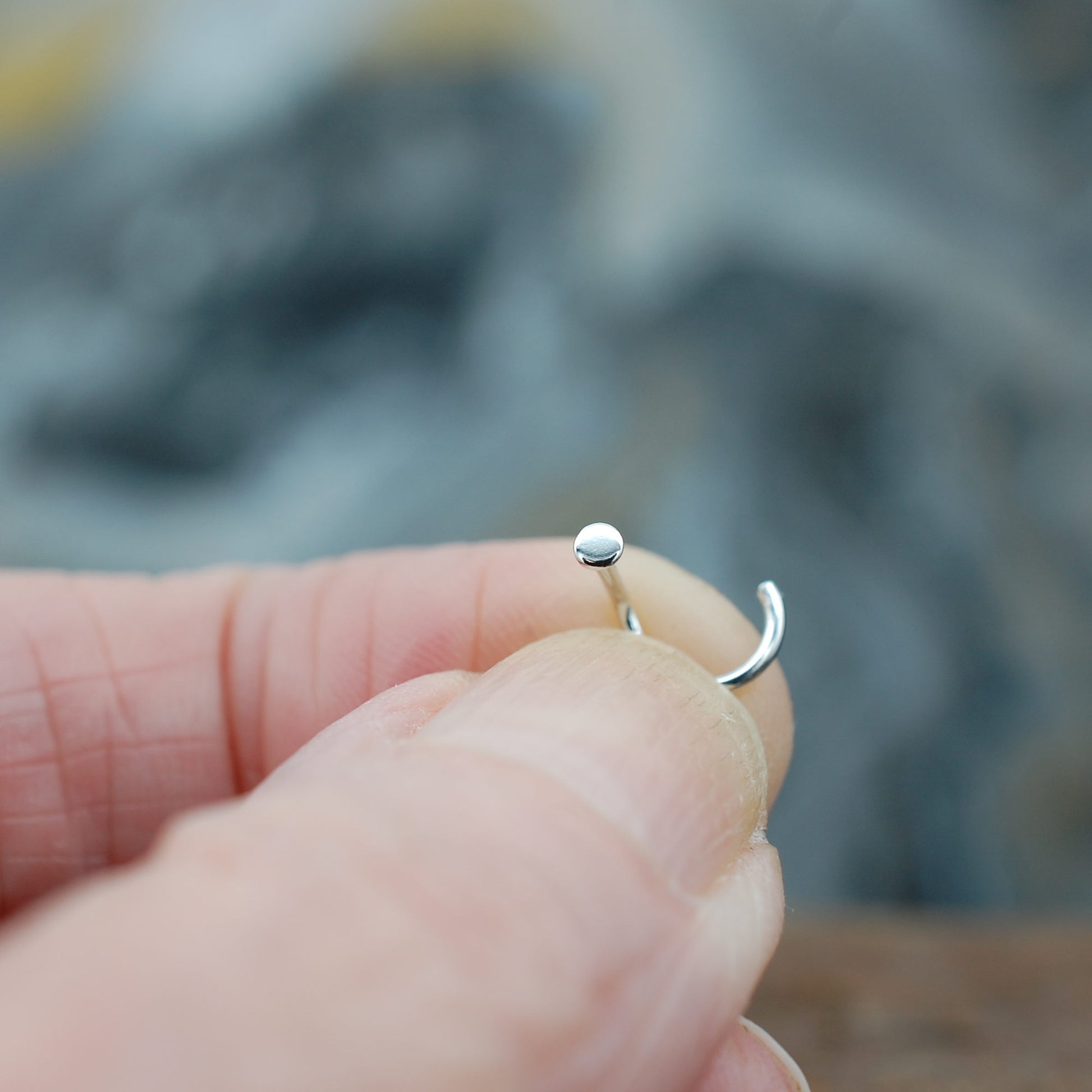 Silver nose ring held between fingers with a blurred natural background