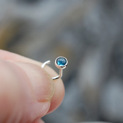Silver nose ring with a blue gemstone held between fingers against a blurred background