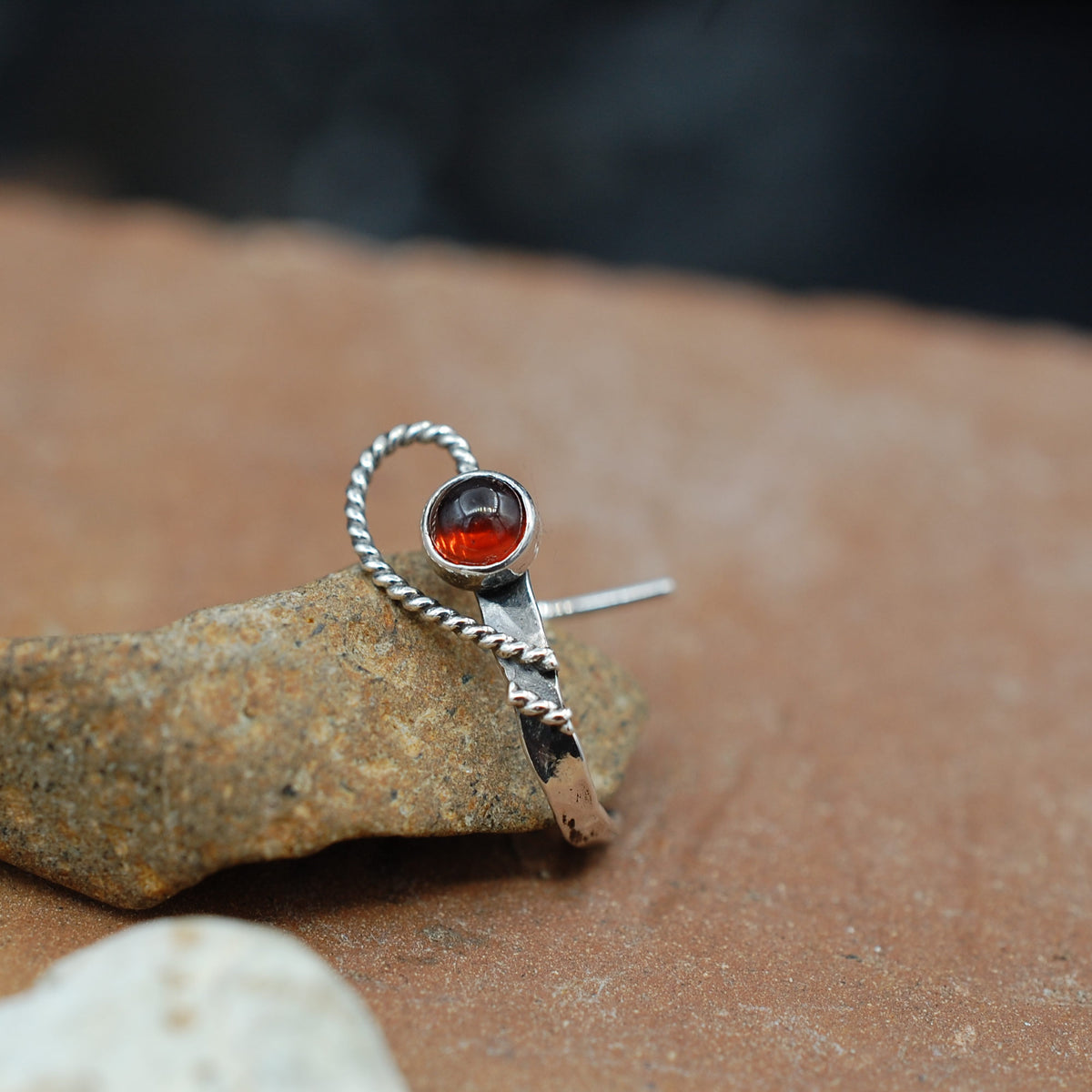 A dappled silver metal nose ring with a twisted wire sweeping open heart design wraps around a garnet gemstone in red.