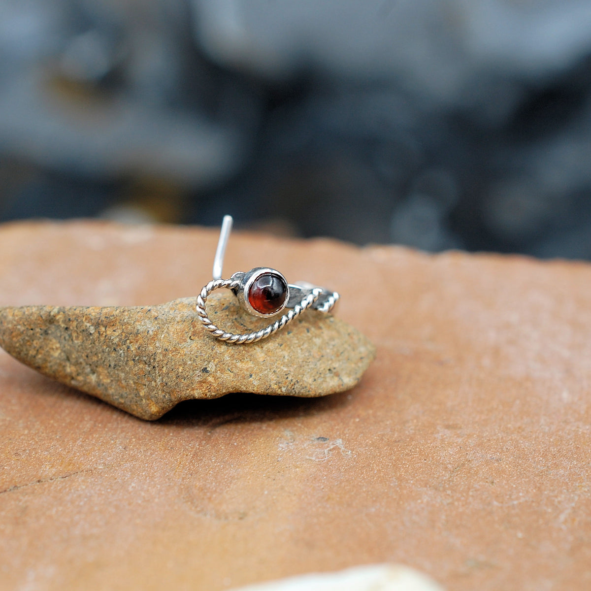 A silver cuff style nose ring laying on its side with the L end pointing up and a garnet gemstone with a twisted swoop are facing front.