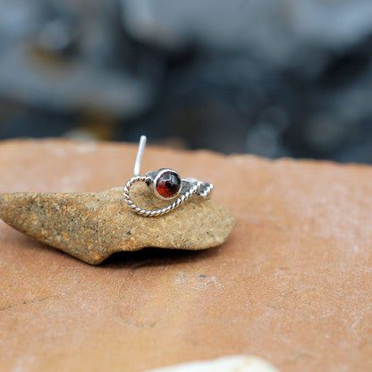 A silver cuff style nose ring laying on its side with the L end pointing up and a garnet gemstone with a twisted swoop are facing front.