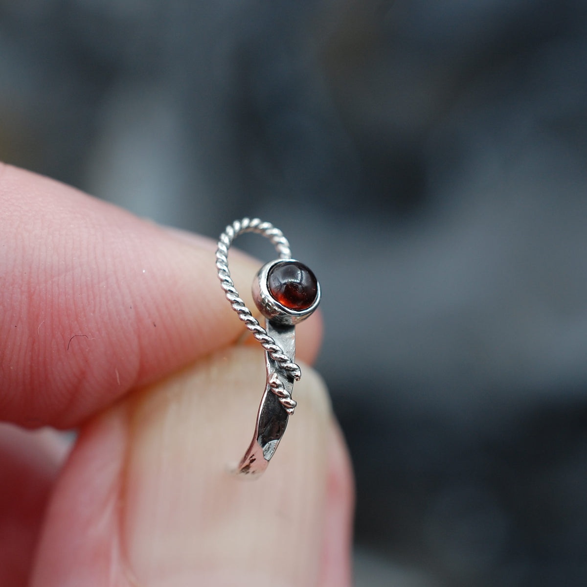 A close up of a hammered nose ring in silver with an open heart design that wraps around a red garnet gemstone. 