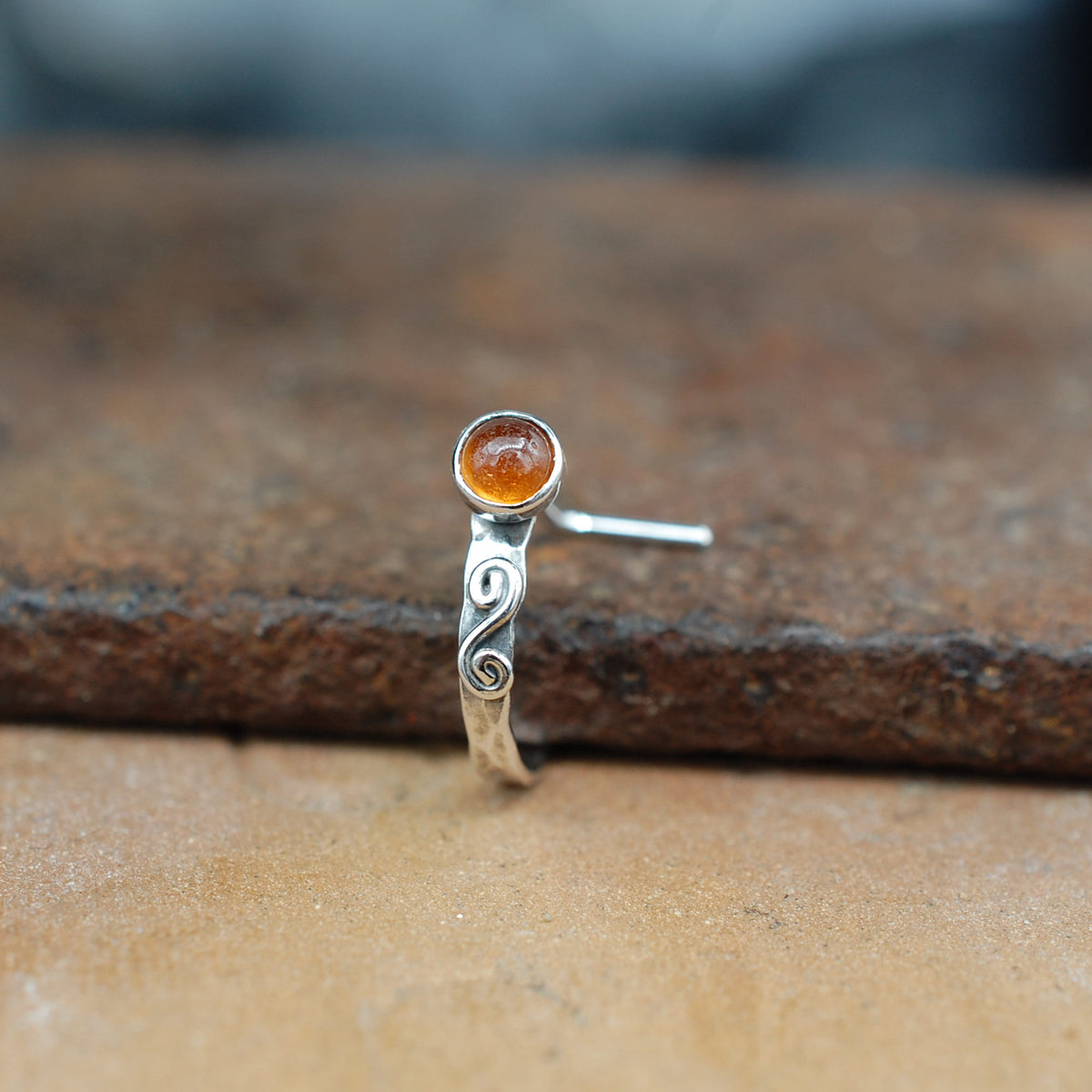 A front facing view of a silver nose ring in a cuff style, embellished with a metal swirl and set with a hessonite garnet gemstone.