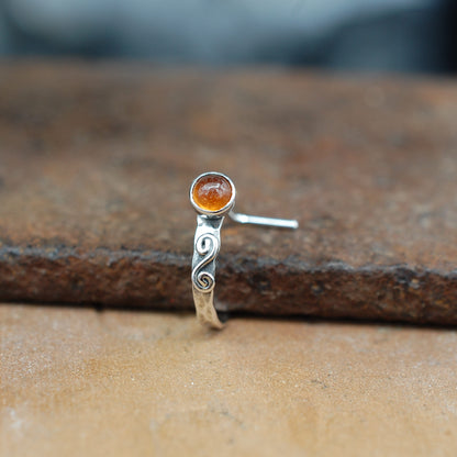 A front facing view of a silver nose ring in a cuff style, embellished with a metal swirl and set with a hessonite garnet gemstone.