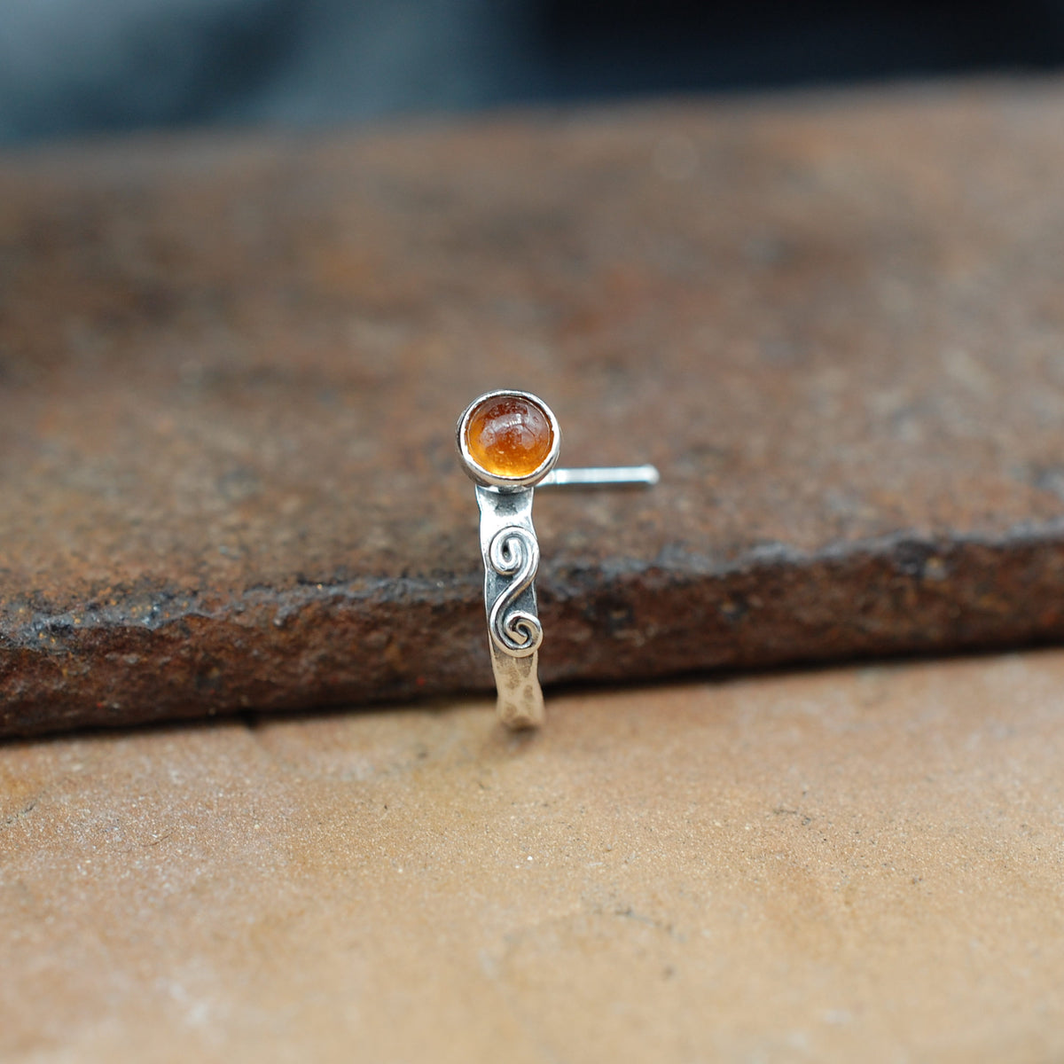 A hessonite garnet gemstone sits on a hammered sterling silver cuff style nose ring and is embellished with a hand twisted swirl.