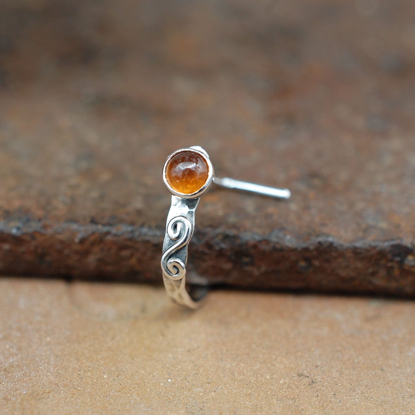 A sterling silver hammered nose ring with a swirl and a round hessonite garnet gemstone, shown on a rustic brown textured surface.
