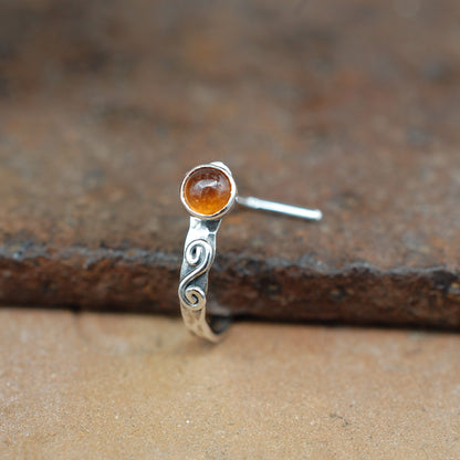 A sterling silver hammered nose ring with a swirl and a round hessonite garnet gemstone, shown on a rustic brown textured surface.