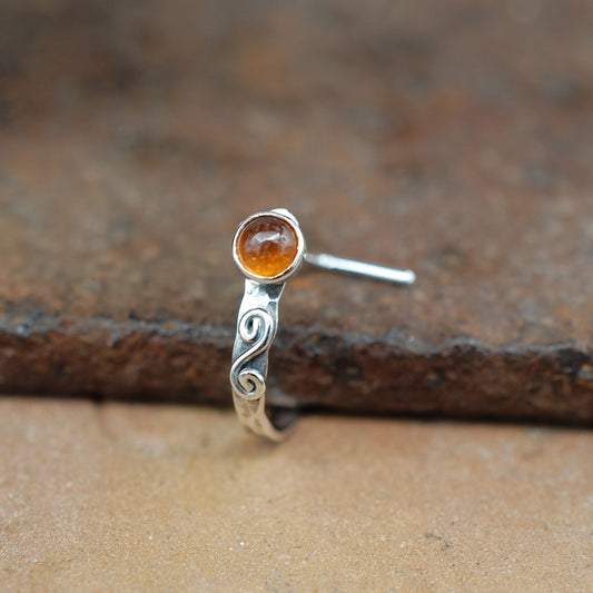 A sterling silver hammered nose ring with a swirl and a round hessonite garnet gemstone, shown on a rustic brown textured surface.