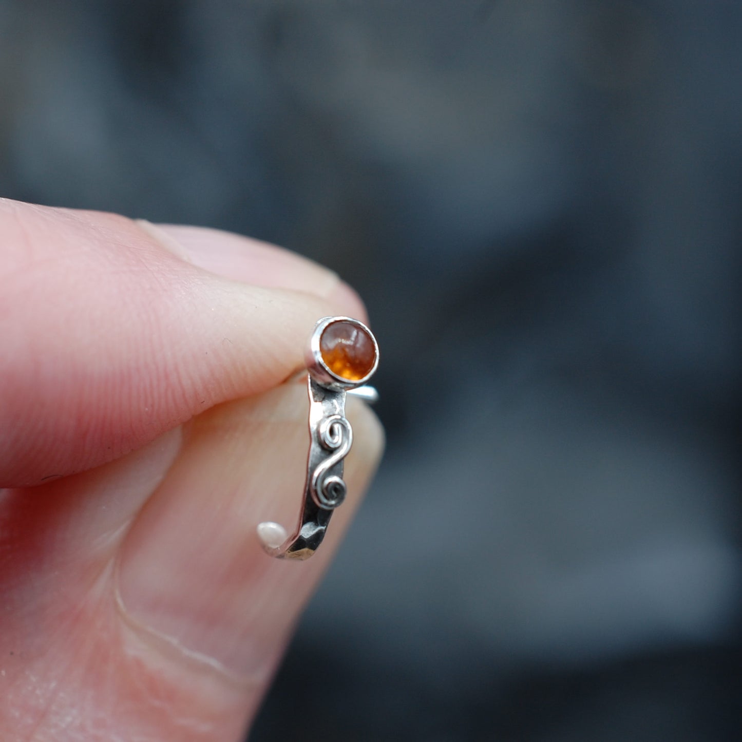 A hammered silver nose cuff set with a hessonite garnet gemstone and embellished with a swirl is held between a thumb and forefinger.