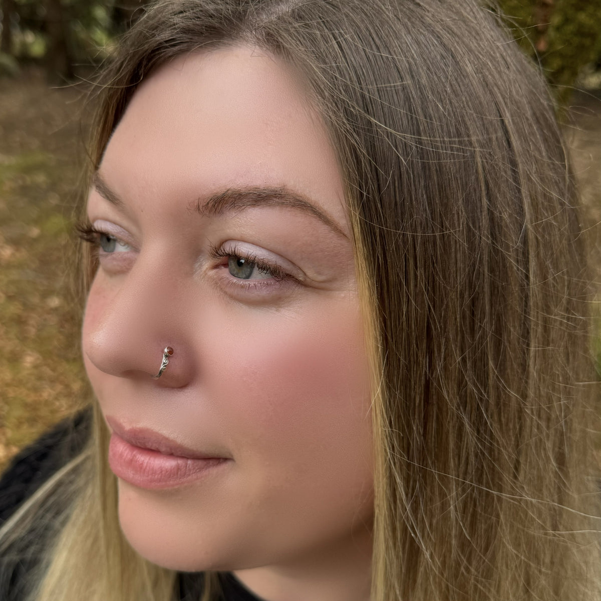 Close-up of a woman outdoors wearing a small silver hammered silver cuff style nose ring with a hessonite gemstone on the left side of her nose.