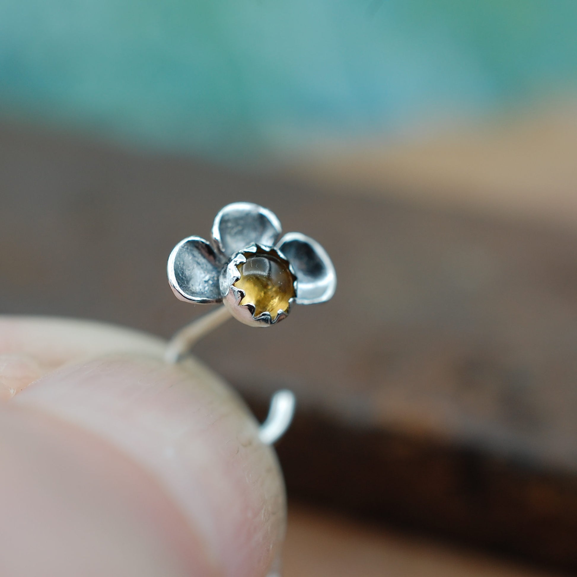 Silver flower-shaped nose stud with a yellow citrine gemstone held between fingers against a blurred background