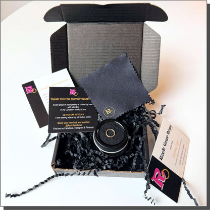 A premium black unboxing experience for Rock Your Nose handcrafted nose jewelry. An open matte-black cardboard mailer box filled with black crinkle paper cleanly showcases a circular black tin container displaying a single gold nose ring on a dark cushion. Elegant black-and-white insert cards are neatly layered on the right, displaying nose jewelry care instructions and a thank you message from the Canadian artisan studio of one. 