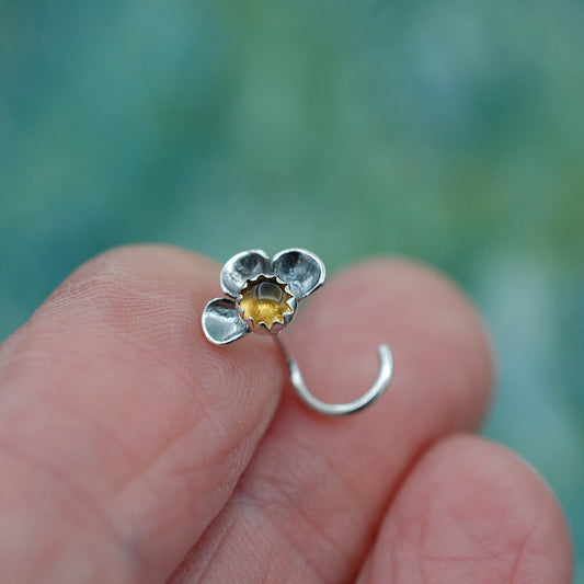 Silver flower-shaped nose stud held between fingers with a blurred green background