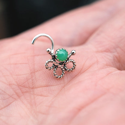 A silver-toned nose hoop ring with a twisted rope bow design and a round green stone, shown resting on a person's palm.