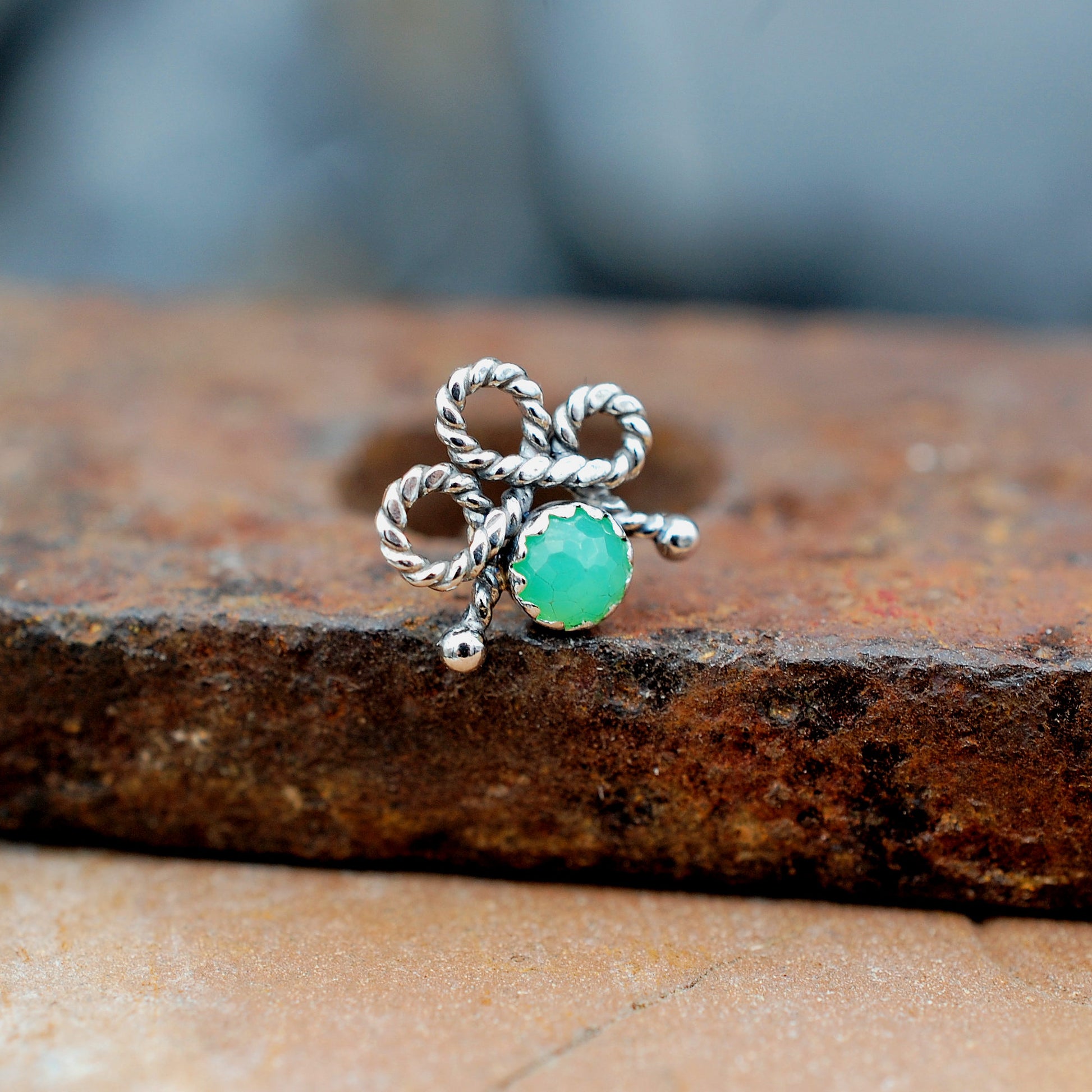 A silver bow-shaped nose stud with a round green stone, resting on a textured rusty metal surface.