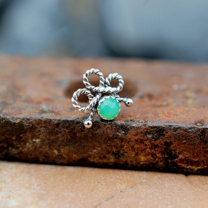 A silver bow-shaped nose stud with a round green stone, resting on a textured rusty metal surface.