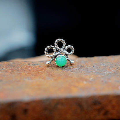 A silver nose stud with a twisted bow design and a round green cabochon gemstone, resting on a textured rusty metal surface.