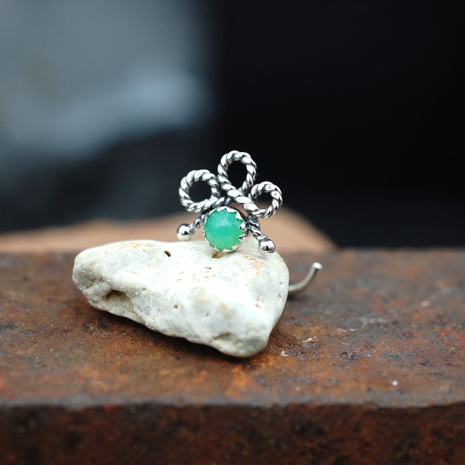 A silver nose stud with a twisted bow design and a round green stone, displayed on a small white rock over a rusty brown surface.
