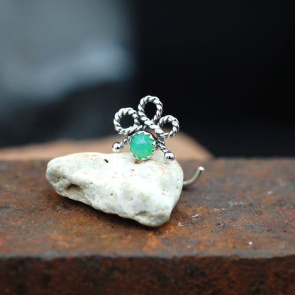 A silver nose stud with a twisted bow design and a round green stone, displayed on a small white rock over a rusty brown surface.