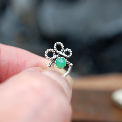 A silver nose stud with a twisted bow design and a round green stone, shown being worn on a person's finger against a blurred outdoor background.
