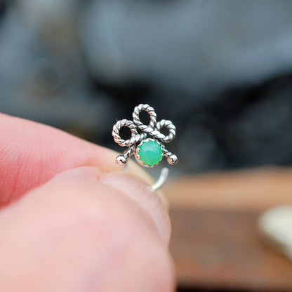 A sterling silver nose stud shaped like a small twisted rope bow with a round green stone in the center, held between fingers against a blurred outdoor background.